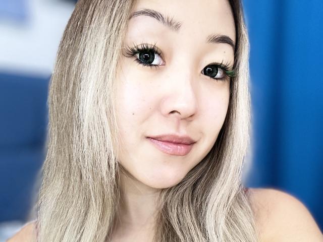 RenyLime has blonde hair, green eyes, long eyelashes, and pink lips. Her light skin is smooth with a small mole on her left cheek. She's smiling slightly.