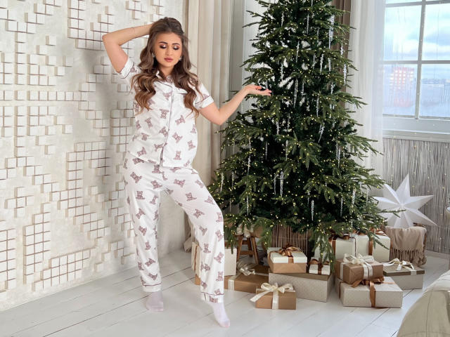 BeataAngel with wavy brown hair, wearing white pajamas with gray reindeer patterns, stands near a Christmas tree, her right hand on her head, left arm out. She's barefoot.
