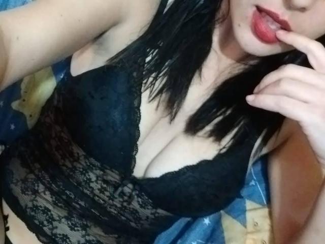 LadyNandas with black lace bra, showing her big boobs, finger in mouth, black hair, pale skin, and red lips, lying down. Her hand is near her face.