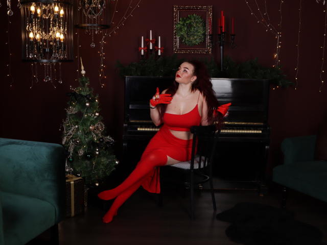 NicolPots in red lingerie, stockings, and gloves, sitting on a chair, showing off her tits, with a Christmas tree and green couch on dark red walls.
