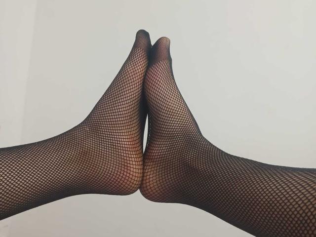 MariahVictoria's feet are crossed at the toes, wearing black fishnet stockings, showing a diamond pattern against a white background.