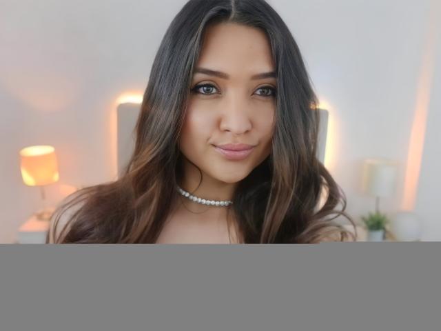 MariahVictoria has long brown hair, a choker, and a cute smile. She's showing her tits in a black top, with warm lighting in the background.