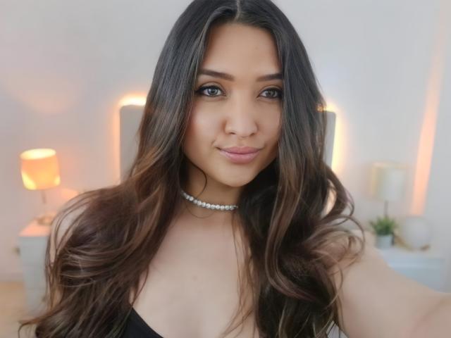 MariahVictoria with long brown wavy hair, wearing a black top and a silver choker, smiling at the camera, showing her cute nose and full lips, with visible breasts.