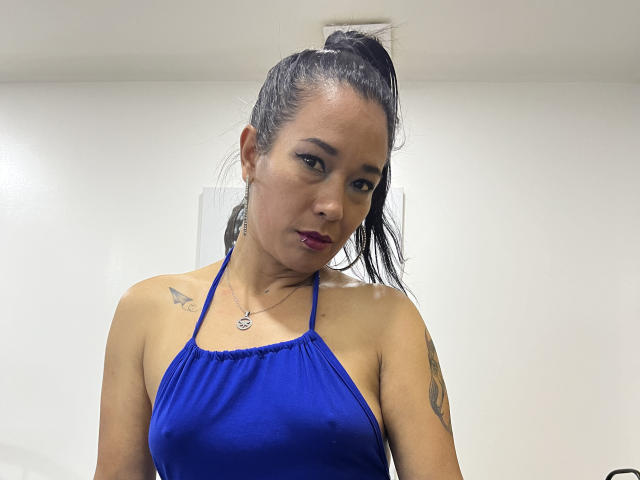 Kylee69 with dark hair in a high ponytail, wearing a blue halter top, showing small tits, tattoos on right shoulder and upper arm, and a necklace.