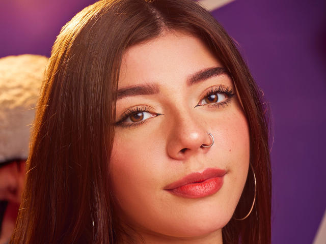 O rosto de Chloe Vertinely, com olhos castanhos, cabelos longos castanhos, um piercing no nariz, lábios rosados, brincos de argola e uma maquiagem discreta, tudo em um ambiente iluminado em tons de roxo. Suas nádegas não estão visíveis..
