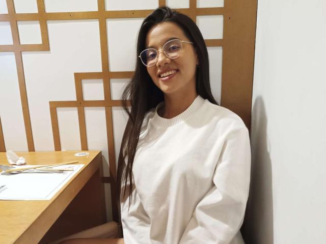 DilanAndMia, con cabello largo y oscuro, gafas y un suéter blanco, sonriendo, sentada, con las manos en el regazo, una mesa de madera a la izquierda, una pared blanca y un panel de madera con diseño detrás..