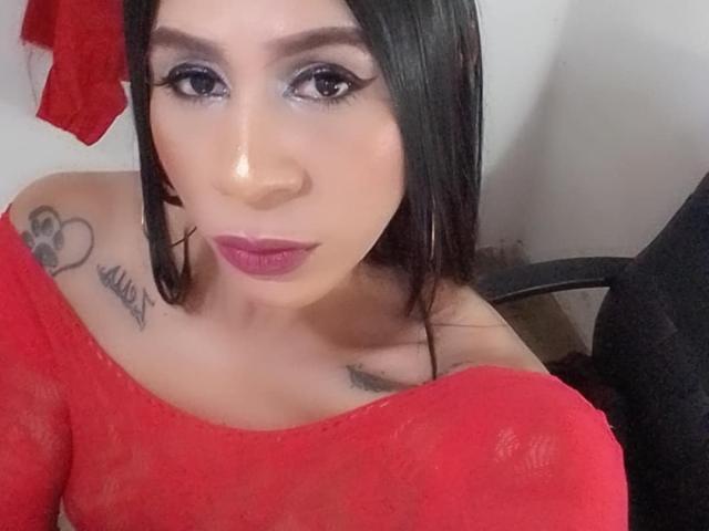 TrayXibi with black hair, red off-shoulder top, purple lipstick, and tattoos on shoulders, looking at camera, showing her tits. She has a nose piercing and hoop earrings.