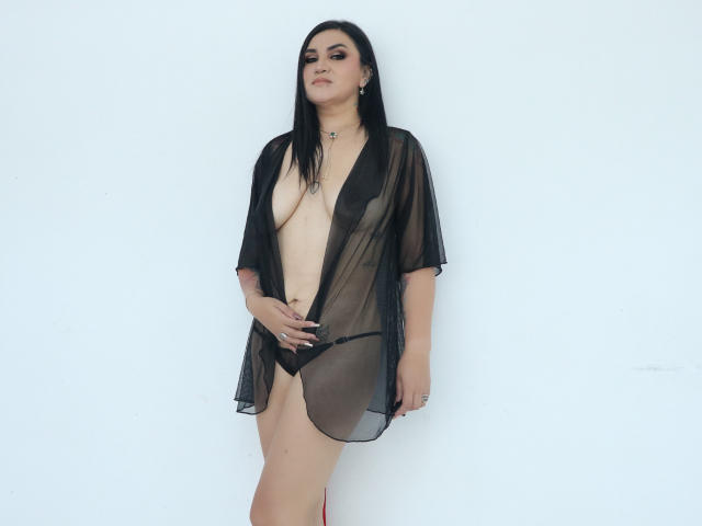 KathyaParsons, dark-haired, standing, wearing sheer black robe, exposing large tits, small black panties, hand on thigh, white background, confident expression, subtle makeup, necklace.