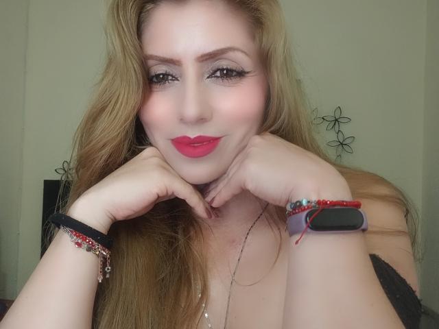 BellaShannon with red lipstick, long blonde hair, and black lace top, resting her chin on her hands, wearing a red beaded bracelet and black smartwatch, smiling slightly.