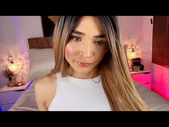 MissAntonellaX with long, straight brown hair, wearing a white halter top, showing her pretty face and pink lips, sitting on a bed with two lit lamps.