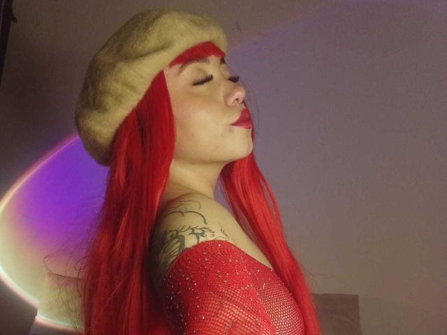 GigiVolkova with red hair, wearing a furry beige hat, red mesh top, and red lipstick, pouting with eyes closed, showing tattooed shoulder, nose ring, and red lips.