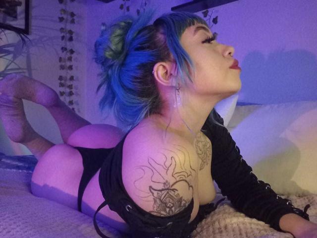 GigiVolkova with blue and green hair, lying on a bed, ass up, black top, tattoos on shoulder and arm, puffy lips, legs in the air, purple lighting.