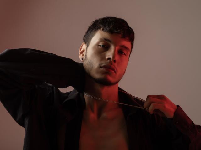 KinkyBoyForYou, senza camicia, con capelli scuri, che tiene una collana a catena sulla spalla, maglietta nera parzialmente aperta, luce rossa sul lato sinistro, espressione seria, leggera barba, scatto in studio..
