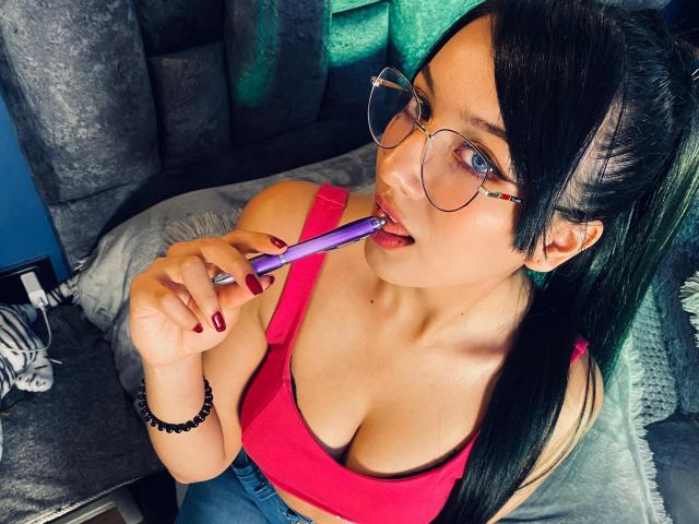 LillyTaylor with black hair in a ponytail, wearing glasses, pink top, and red nails, sucking a purple dildo, showing her big tits. She's on a gray couch.