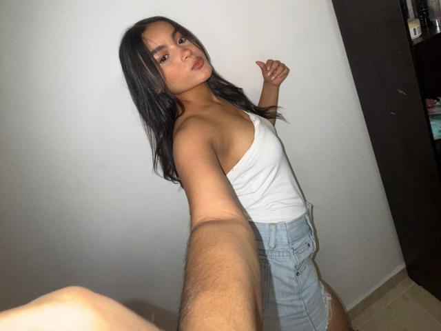 MiiaPalmer with long black hair, wearing a white tank top and light blue denim shorts, taking a selfie, showing her small tits and slim ass, against a white wall.