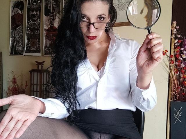 MiniBeast with long black hair, wearing glasses, white button-up, black skirt, and sheer pantyhose, holding a magnifying glass, looking at camera with red lips, showing small tits.