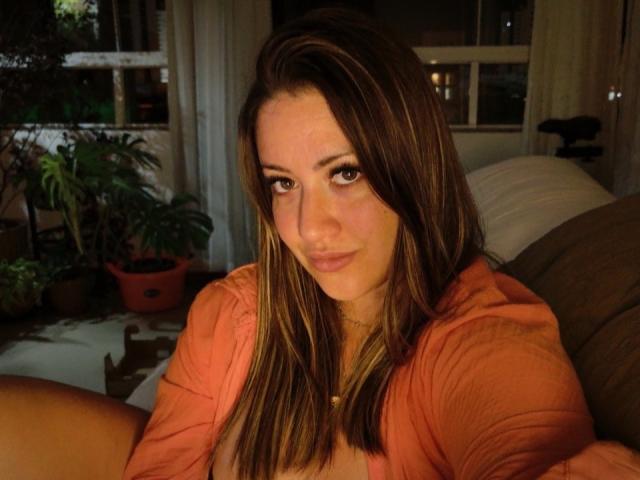 Anyellen with brown hair, wearing an orange shirt, sitting on a couch, looking at the camera with a slight smile, her tits partially visible, in a dimly lit room.