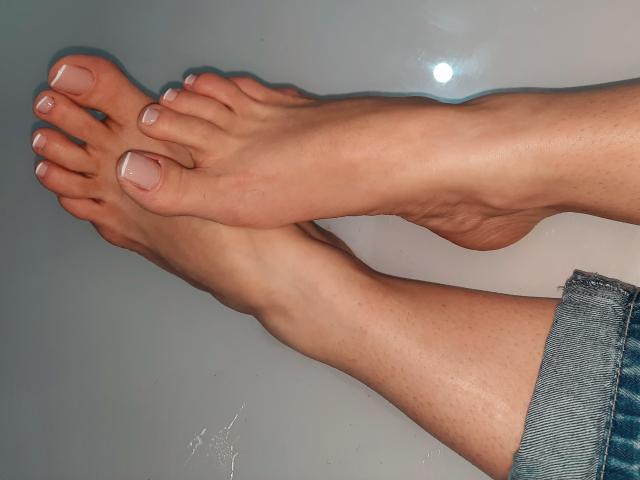 Anyellen's feet, showing tan skin with white nail polish on toes, crossed at the ankles, and wearing blue denim jeans. Her bare legs and feet are visible.