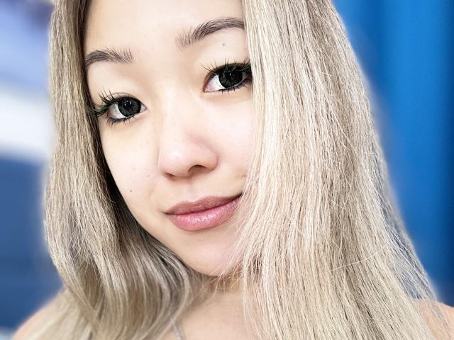 RenyLime has blonde hair, full lips, and long eyelashes, smiling softly at the camera. Her cute face and blue eyes are front and center. She's looking very sexy.