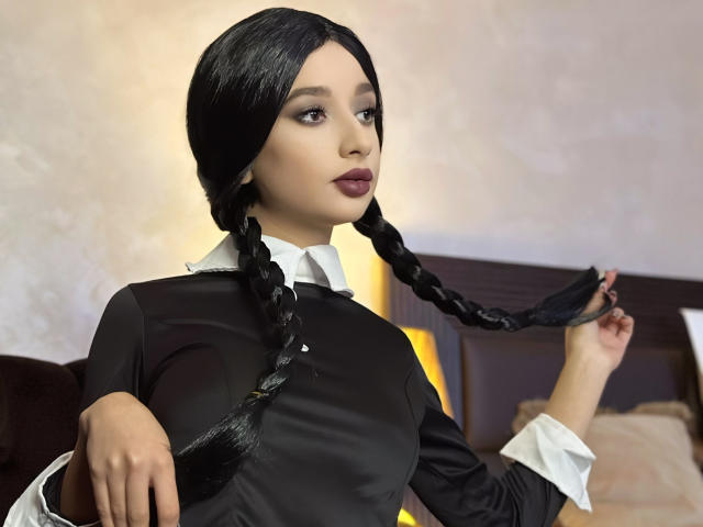 Odette Benson, con due trecce nere, indossa un abito di raso nero con polsini bianchi, tiene una treccia, ha il rossetto scuro e un'espressione seria. Seno, trecce, abito di raso..