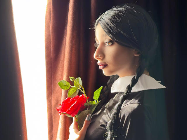 Odette Benson, con trecce scure, che tiene una rosa rossa, con la luce del sole sul viso, vestita con un abito bianco e nero, con una tenda marrone sullo sfondo, che guarda di lato con un'espressione calma..