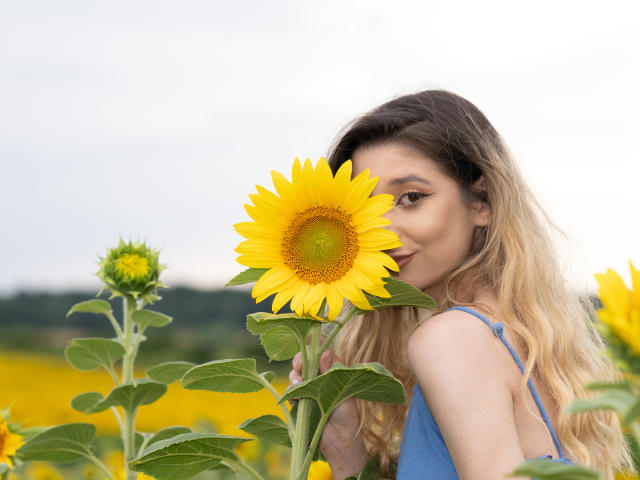 OdetteBenson houdt een enorme gele zonnebloem vast, die gedeeltelijk haar gezicht bedekt, met lang blond haar, en draagt een blauwe top, in een veld met zonnebloemen, waarbij haar borsten nauwelijks zichtbaar zijn..