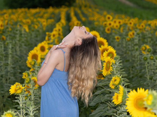 OdetteBenson with long blonde hair in a blue dress, head tilted back, eyes closed, touching her neck, surrounded by tall sunflowers in a field. Her tits are slightly visible.