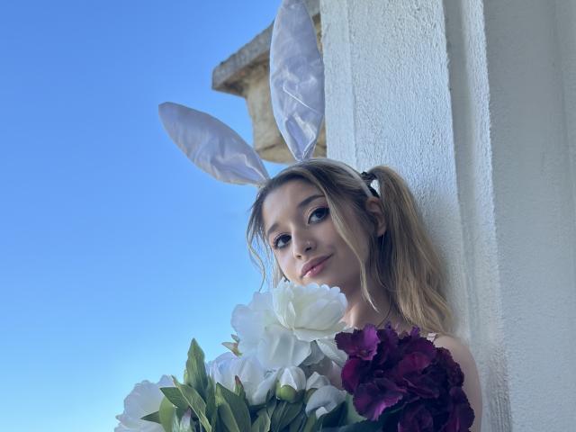 OdetteBenson with puffy white bunny ears, holding a bouquet of white and purple flowers, has blonde pigtails, and a cute, slightly sexy face, against a white wall.