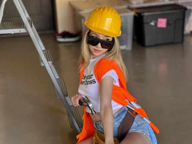OdetteBenson, wearing a yellow hard hat, black sunglasses, orange vest, white shirt, and denim shorts, holds a ladder with a tool, showing a sexy, confident, construction worker look.