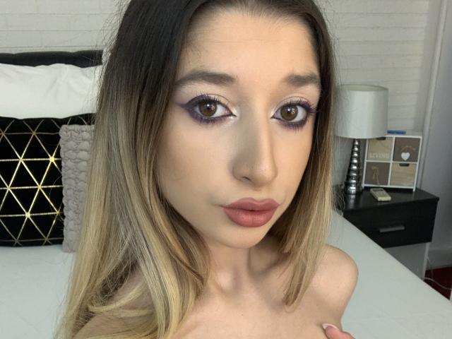 OdetteBenson has blonde ombre hair, brown eyes with purple eyeshadow, and pink lipstick. She's topless, showing her tits, in a bedroom with a black and white bed.