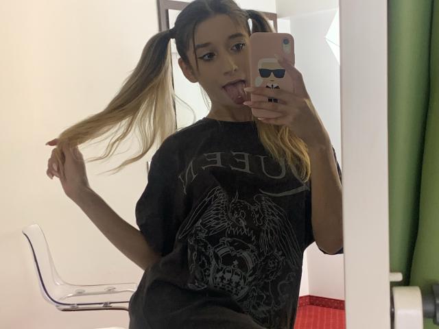 OdetteBenson with pigtails, tongue out, holding hair, wearing a black "AC/DC" shirt, mirror selfie, phone with cute face sticker, clear chair, green curtain in background.