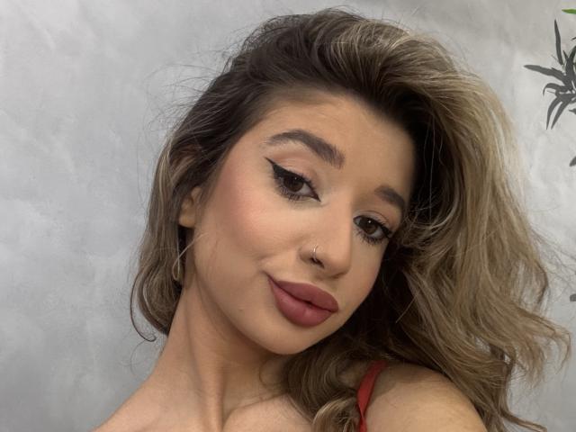 OdetteBenson with wavy blonde hair, brown eyes, winged eyeliner, red lipstick, nose ring, and hoop earrings, smiling at camera, wearing red strap, looking sexy.