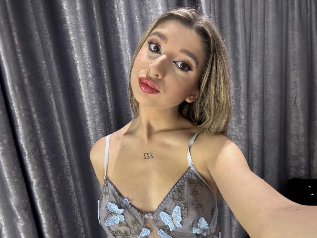 OdetteBenson with blonde hair, wearing a sheer blue butterfly bra, showing small tits, tattoo "SS" on chest, red lips, and brown eyes, against gray curtains.
