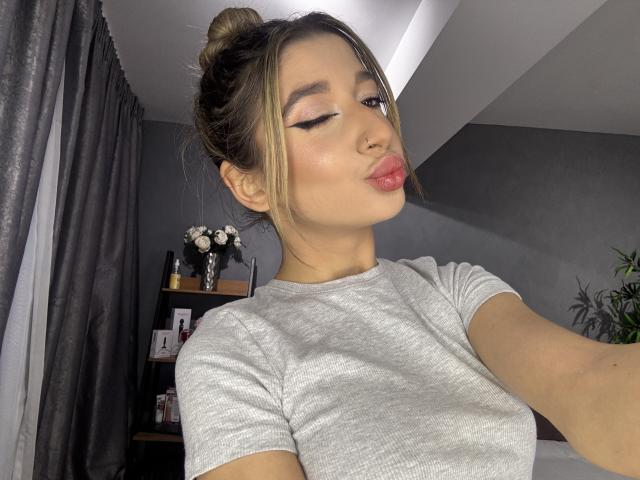 OdetteBenson with her tits in a grey tee, pouty lips, and eyeliner, has her blonde hair in a bun, winking, and nose pierced, in a brightly lit room.