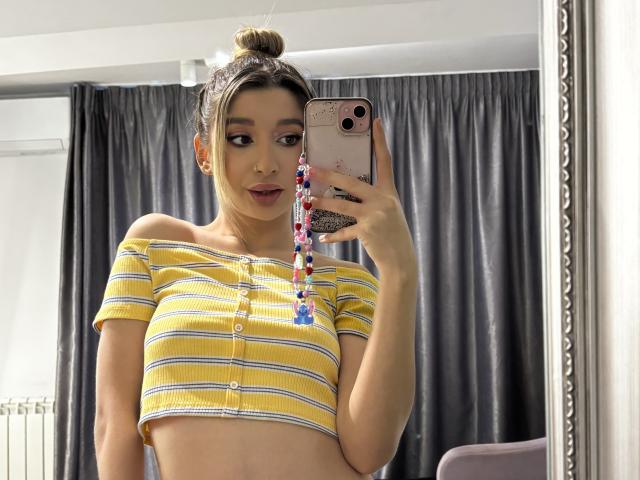OdetteBenson with her tits out, wearing a yellow off-shoulder crop top, holding a phone with a colorful strap, her hair in a bun, taking a mirror selfie.