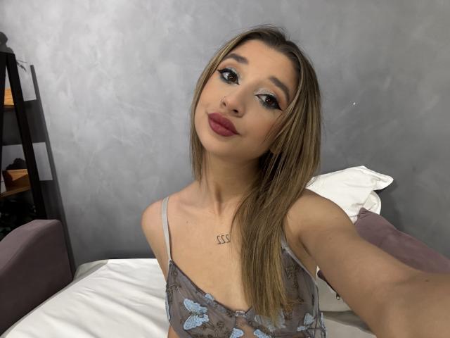 OdetteBenson has long blonde hair, wearing a sheer blue butterfly top, showing small tits, with red lipstick and a nose ring, taking a selfie in a bedroom.