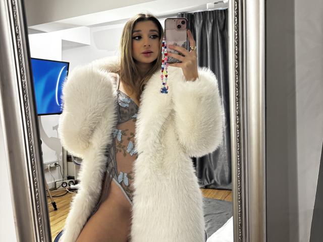 OdetteBenson with long brown hair, wearing a fluffy white coat, sheer grey lingerie with blue floral patterns, holding a pink phone, showing her tits and pussy in a mirror.