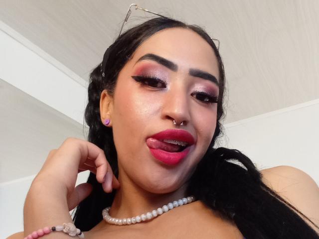MichellDiaz, avec des cheveux foncés, de l'eye-liner rose, des sourcils épais, un anneau au nez, du rouge à lèvres rouge, la langue tirée, un collier de perles et des boucles d'oreilles, regardant vers le bas avec la main près de la bouche..