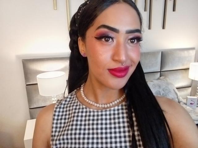MichellDiaz ha lunghi capelli neri, ombretto rosa, rossetto rosso, un piercing al naso e una collana di perle, indossa una maglietta a scacchi bianchi e neri, ed è molto carina e attraente..