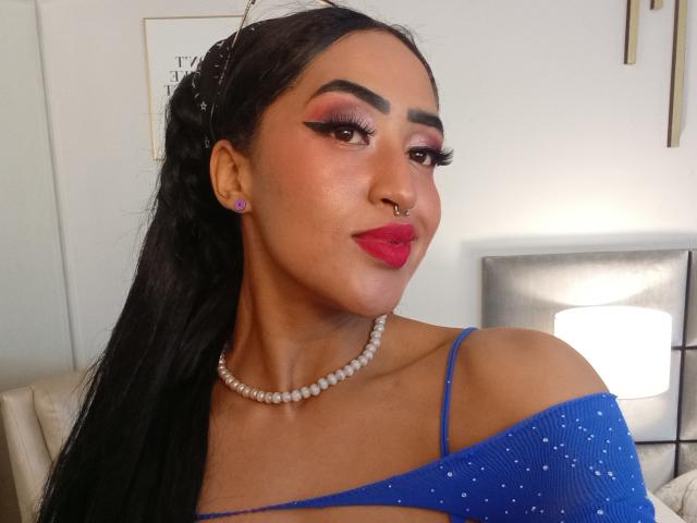 MichellDiaz ha i capelli scuri, rossetto rosso, sopracciglia marcate e una collana di perle. Indossa una maglietta blu con le spalline scoperte e con pois bianchi, e ha un piercing al naso..