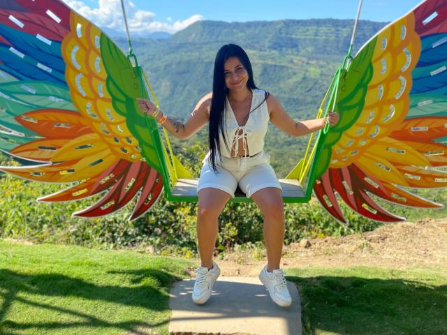HikariChloe with long black hair, white sleeveless top, and white shorts, sitting on a colorful swing with rainbow wings, wearing white sneakers, smiling outdoors.