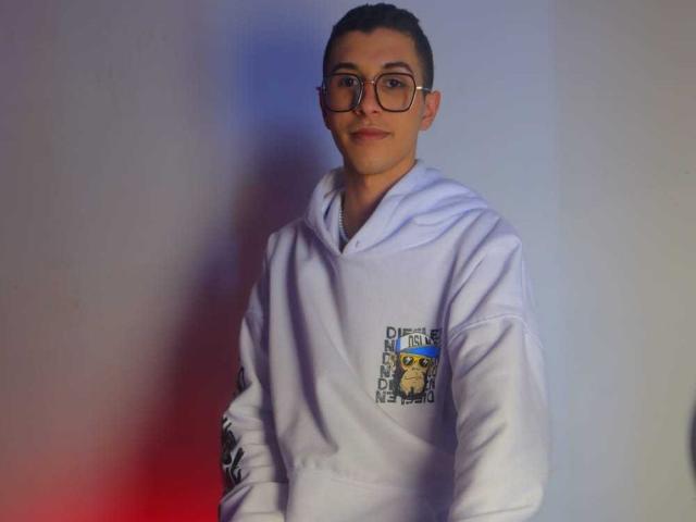 ThomasTwink69, nerdy dude with glasses, short brown hair, white hoodie, small yellow cartoon on chest, slightly smiling, standing against gradient blue and red wall.