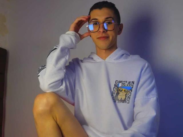 ThomasTwink69 mit Brille, kurzem, dunklen Haar, weißer Kapuzenjacke, sitzt mit angezogenem Knie, Hand am Schläfenbein, leichtes Lächeln, blickt in die Kamera, hellblaue Wand, lässig, süß..