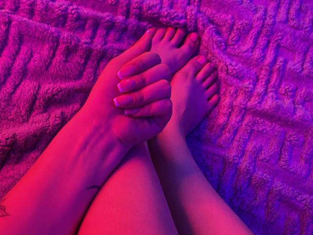 Olieri's feet with hands on top, pink toenails, and a small tattoo on the ankle, illuminated by purple and pink lighting, resting on a textured pink blanket.
