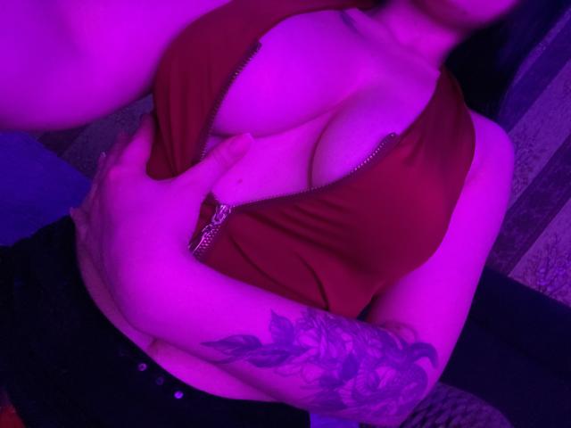 Olieri's big tits are exposed in a red zip-up top, with a hand covering her chest and a tattooed arm visible, under purple lighting.