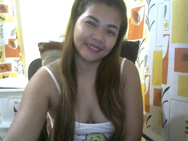 SweetJolieNikki with long brown hair, smiling, wearing a white tank top showing her small tits, sitting in a chair, in a brightly colored room.