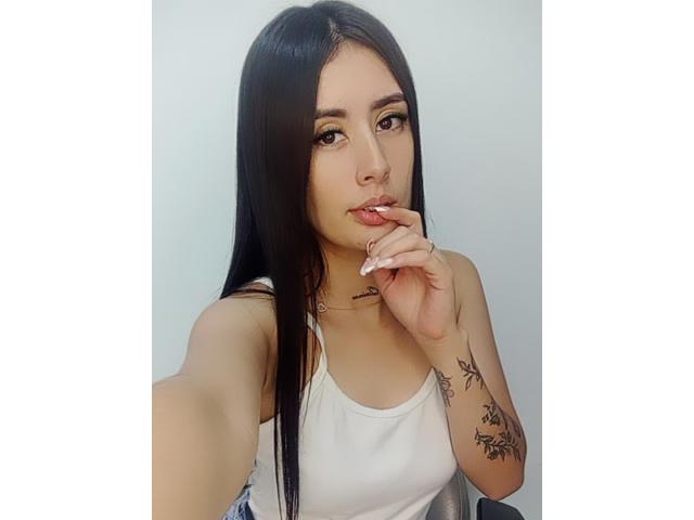 AlisonKweel tiene el pelo largo y negro, ojos marrones y un piercing en la nariz. Lleva una camiseta blanca sin mangas, se está mordiendo el dedo y tiene tatuajes en el brazo..