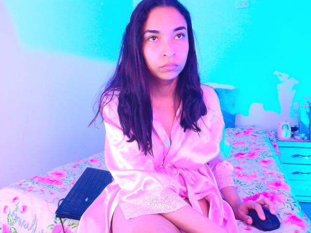 SarahyJhones in a pink silk robe, sitting on a bed with floral sheets, has long dark hair, a cute face, and a small, subtle ass, looking at the camera.