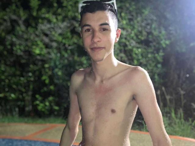 ThomasTwink69, shirtless, with water droplets on his skin, has short dark hair, a white hairdryer on his head, and a slight smile, standing by a pool at night.