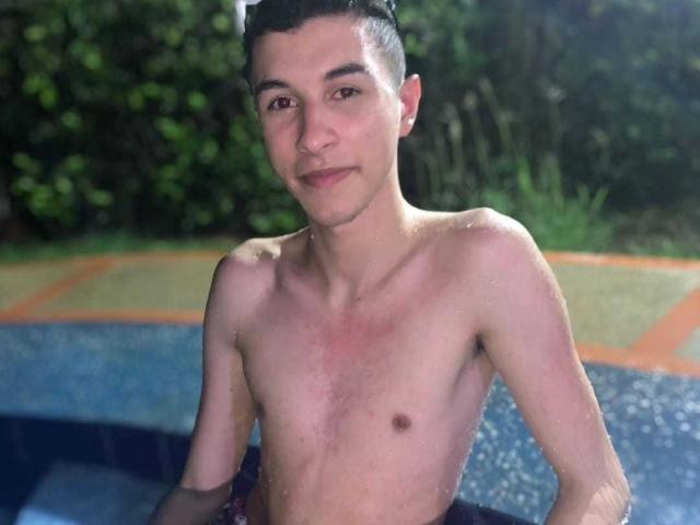 ThomasTwink69, torse nu, avec la peau mouillée, se tient dans une piscine, il a une silhouette mince, des cheveux foncés et un léger sourire, laissant apparaître une petite marque rouge sur sa poitrine..